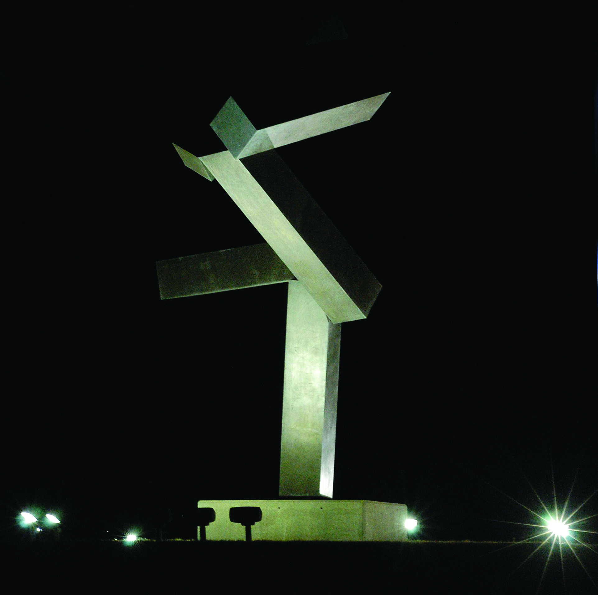 Nighttime view of a large abstract metal sculpture with intersecting beams, dramatically lit from below against a black sky.