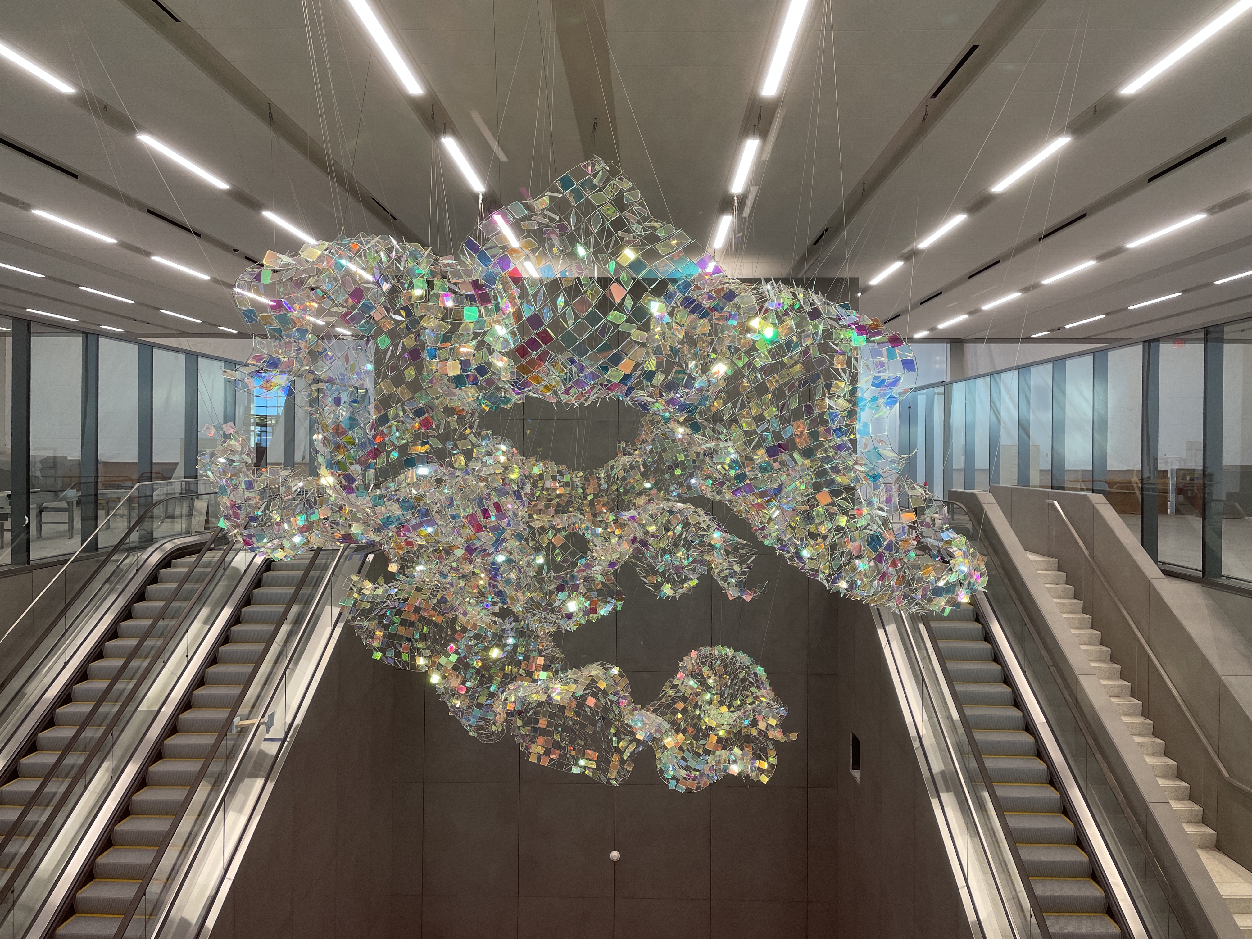 Suspended sculpture of iridescent, net-like mesh glimmering above escalators at Kansas City International Airport.