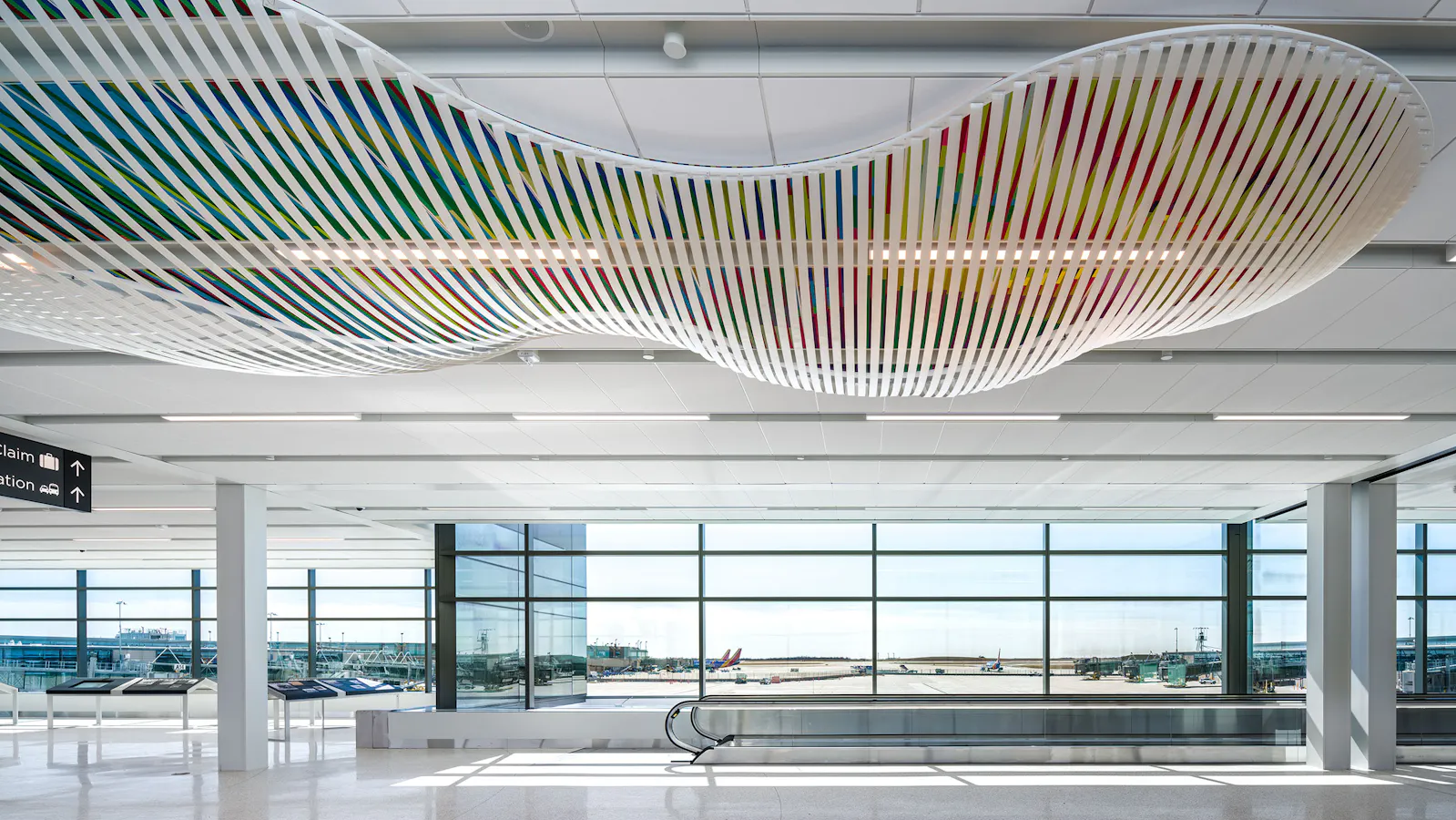 Colorful ceiling sculpture with undulating white lattice structure and diagonal rainbow bands suspended above a wide airport walkway with floor-to-ceiling windows and moving sidewalks.