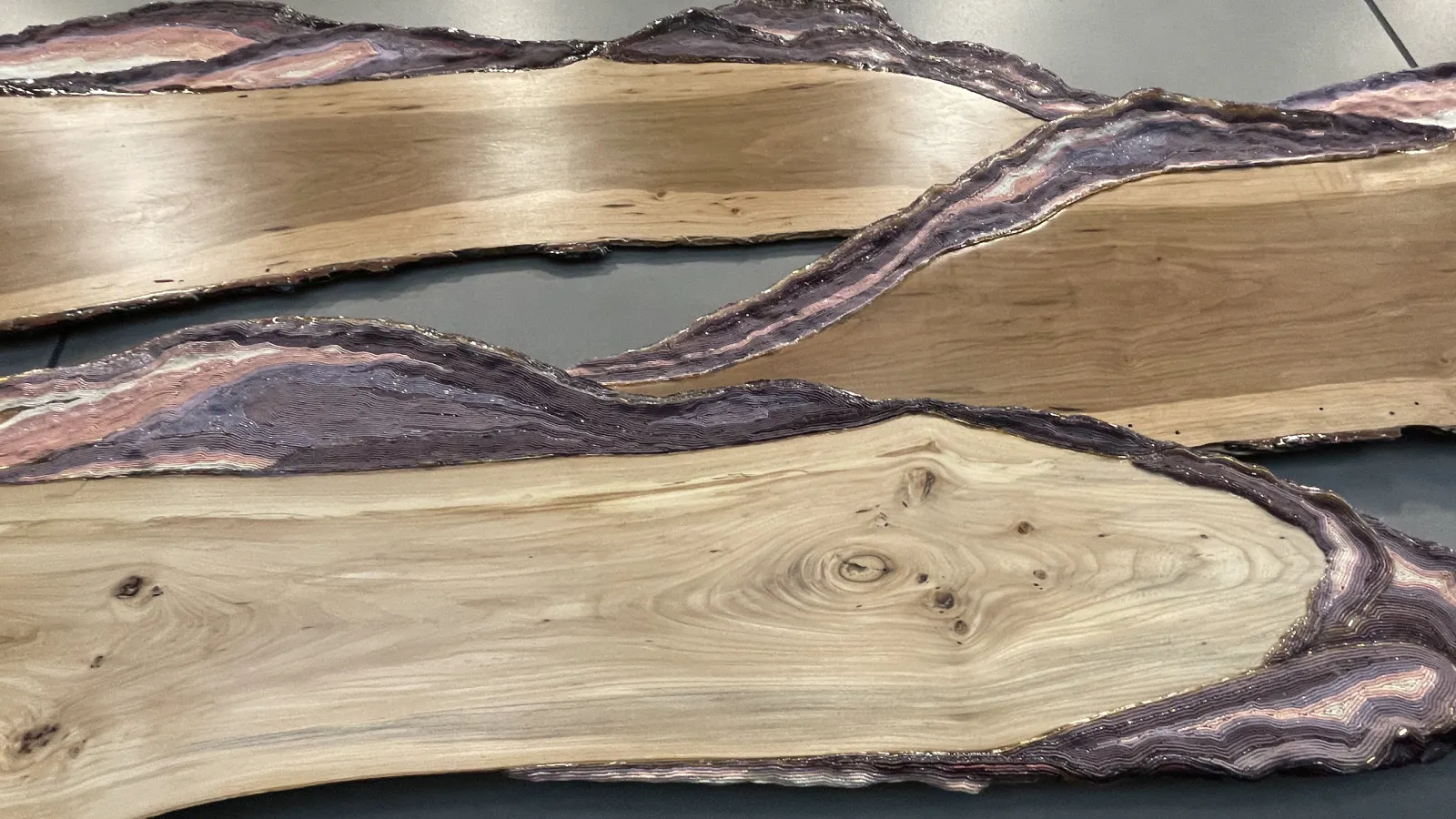 Close-up of sculptural wooden slabs with swirling grain patterns and textured, layered purple and pink resin edges resembling topographic lines or geological formations.