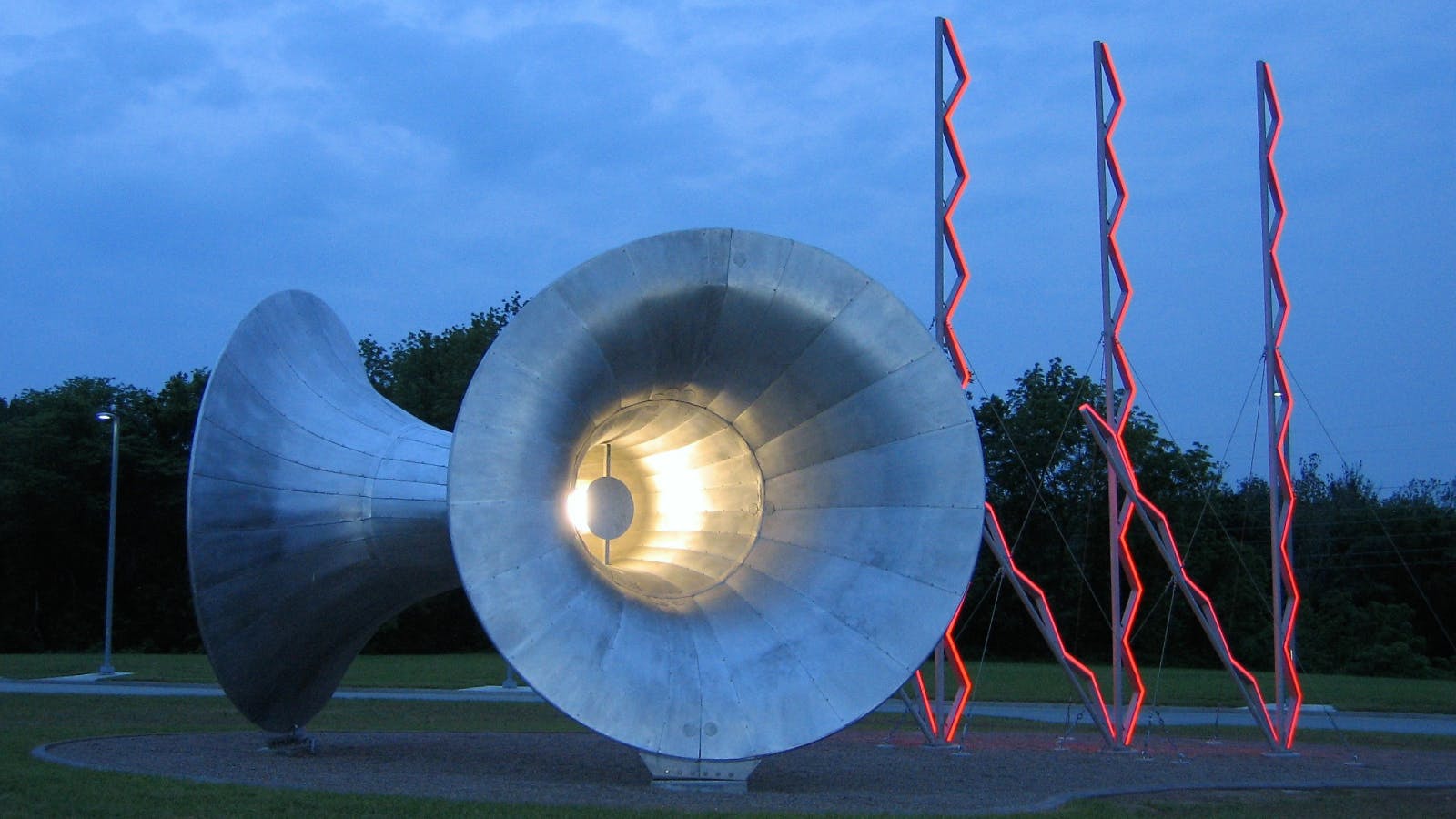 Evening view of a large metallic horn-shaped sculpture lit from within, with red zigzag light structures standing behind it on a grassy field.