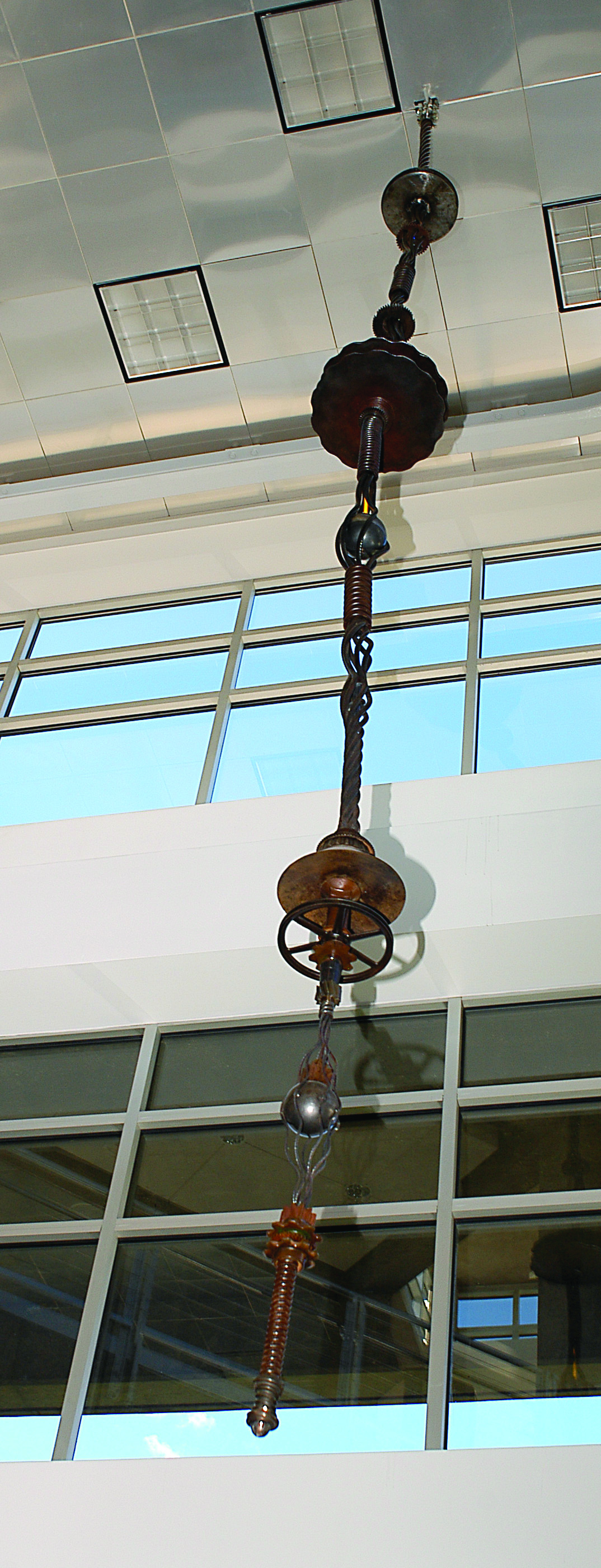 Vertical hanging sculpture composed of various metal and mechanical elements, suspended from a high ceiling in front of large windows with sky views.