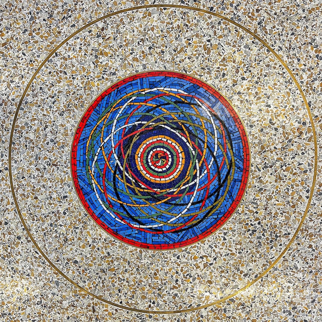 Colorful circular mosaic embedded in a terrazzo floor, featuring layered rings and swirling lines in red, blue, and gold tones.