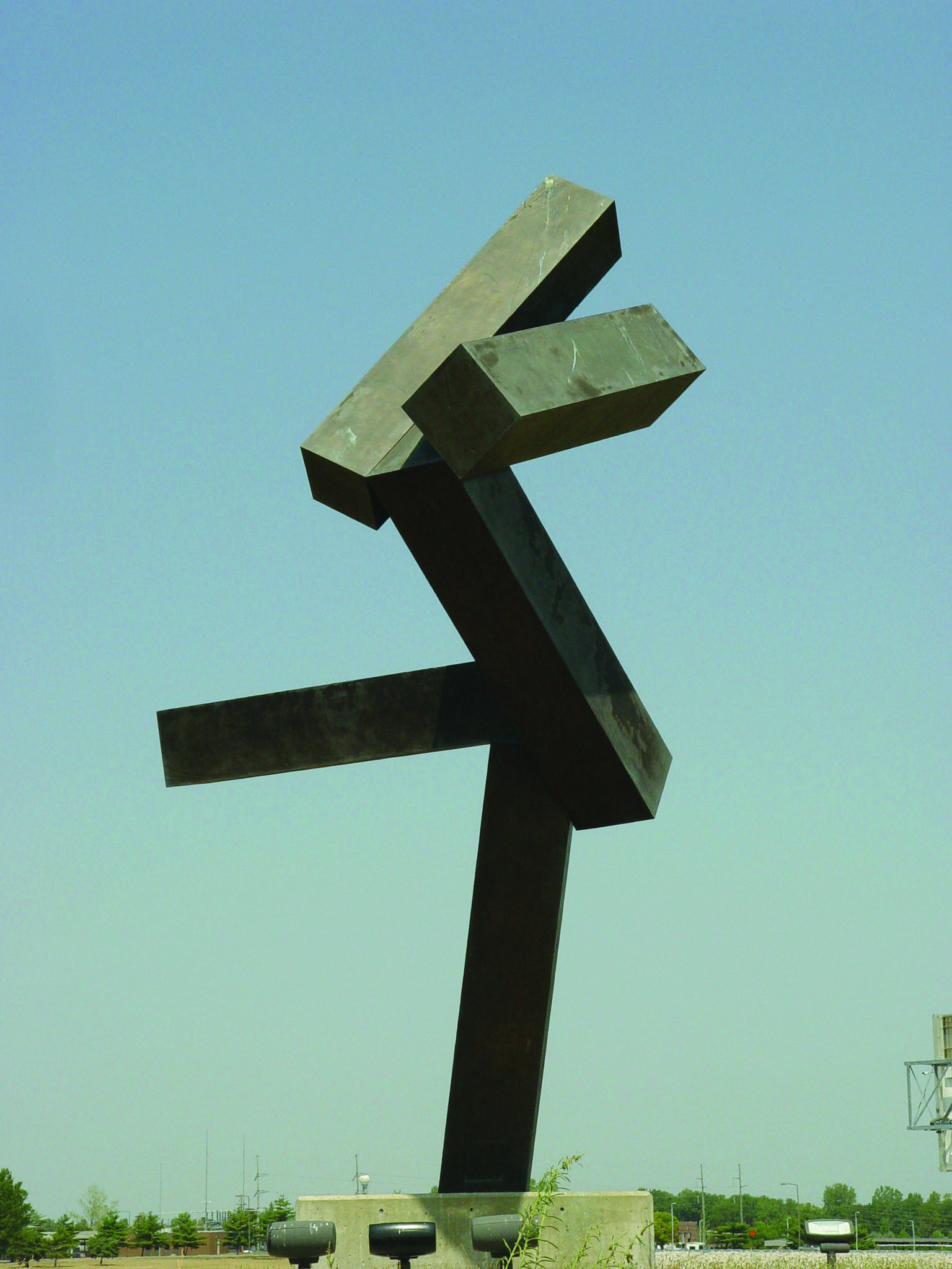 Tall abstract metal sculpture composed of large intersecting beams, angled dynamically against a clear blue sky.