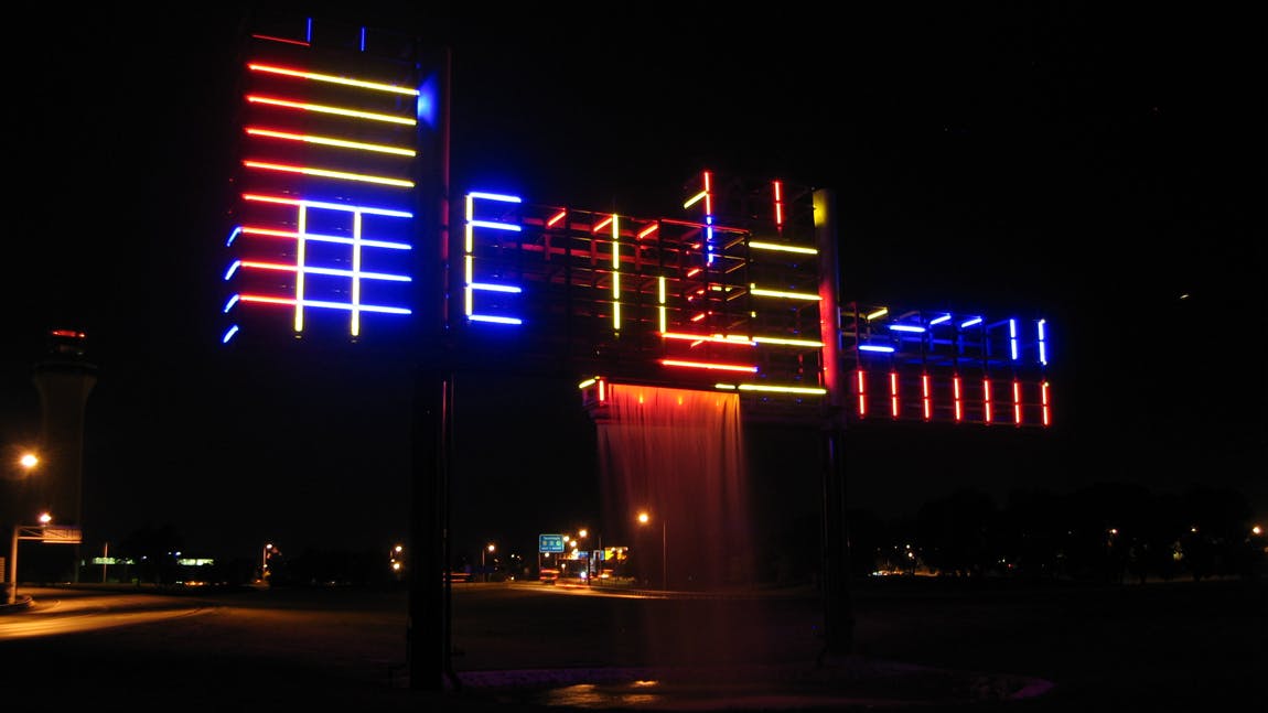 Colorfully lit fountain sculpture at night with red, blue, and yellow neon-style lights above cascading water, set against a dark sky.