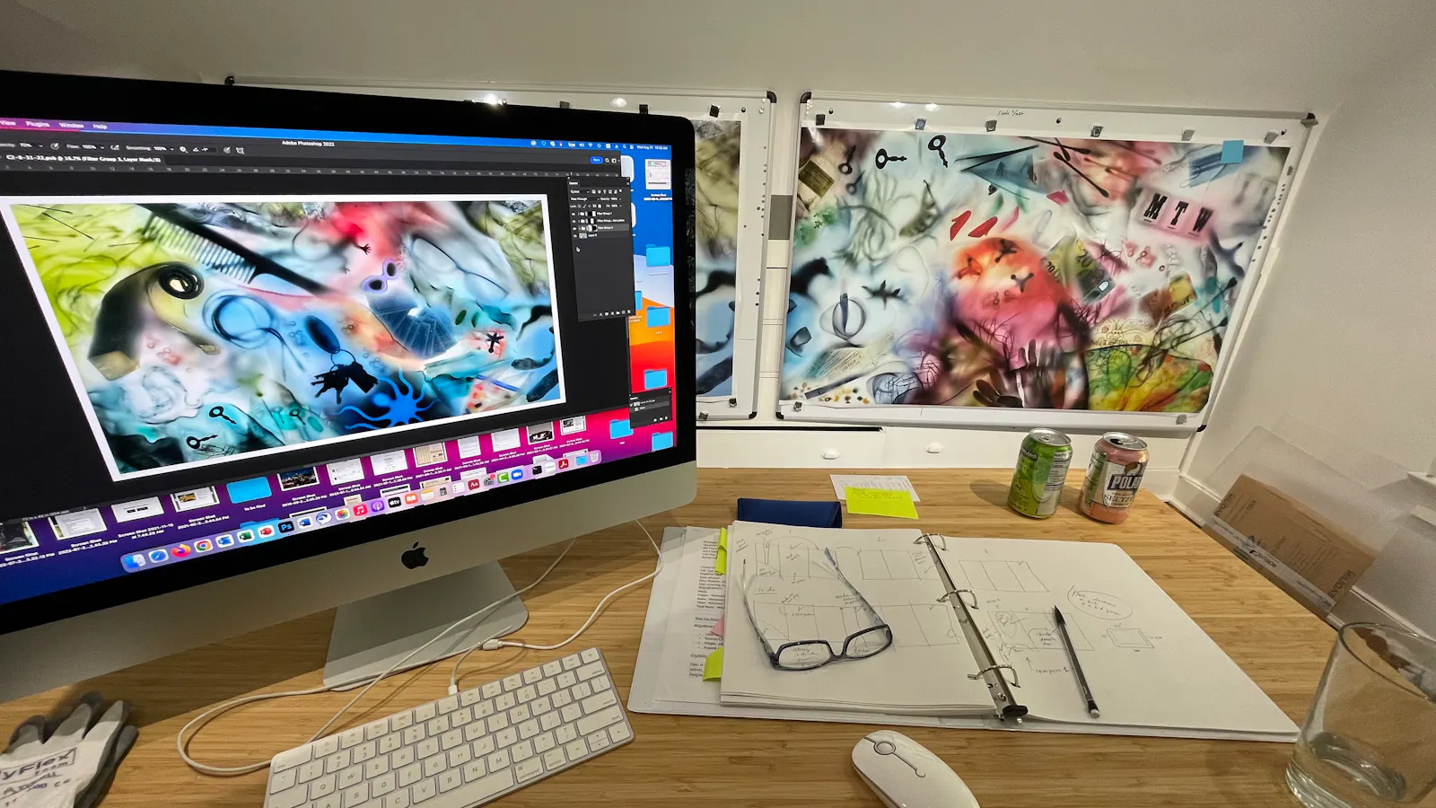 Artist’s workspace showing a desktop computer displaying digital artwork, with printed versions of the same abstract collage pinned behind it and sketch materials spread across the desk.