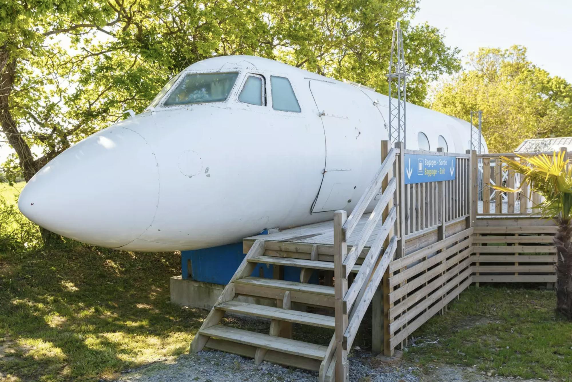 Den Eingang zum Flugzeug-Airbnb erreicht man über eine Holztreppe. Um das Flugzeug herum befinden sich Bäume und ein Feld. 