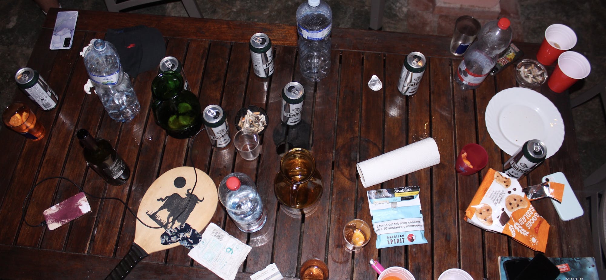 Na drewnianym stole leży różnego rodzaju rzeczy, od puszek z piwem, przez ciasteczka, aż po rakiety do gry w piłkę, papierosy, wodę i Aperol.