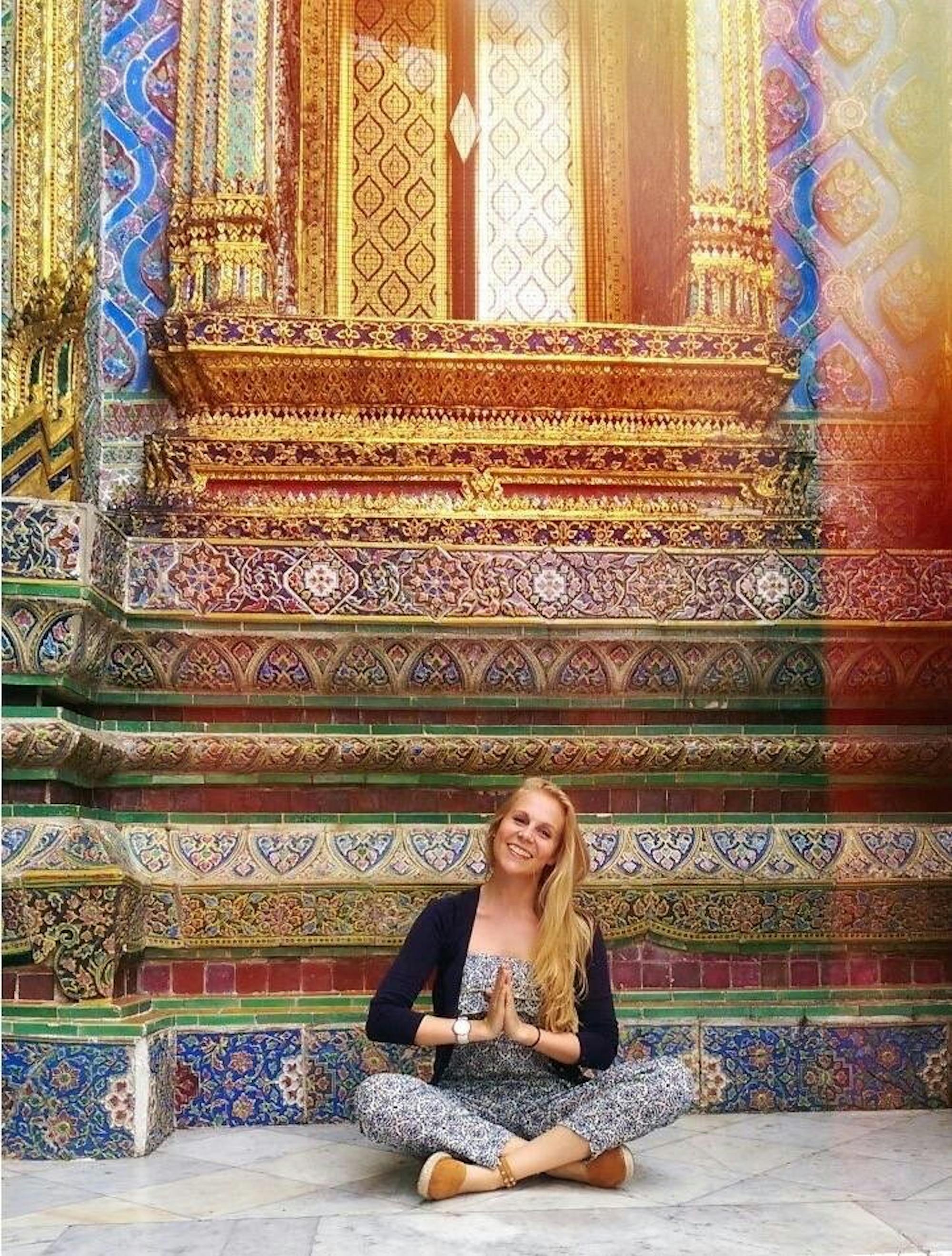 Eine junge Frau (Meike) sitzt im Schneidersitz in meditierender Position in einem Tempel. Hinter ihr sind farbenfrohe Malereien zu sehen. 