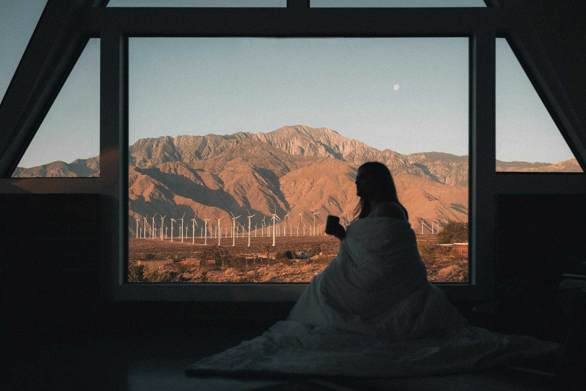Eine Frau sitzt mit ihrer Bettdecke eingekuschelt am Fenster und genießt ihren Morgenkaffee mit einer wundervollen Aussicht auf die Windparks in den USA.  