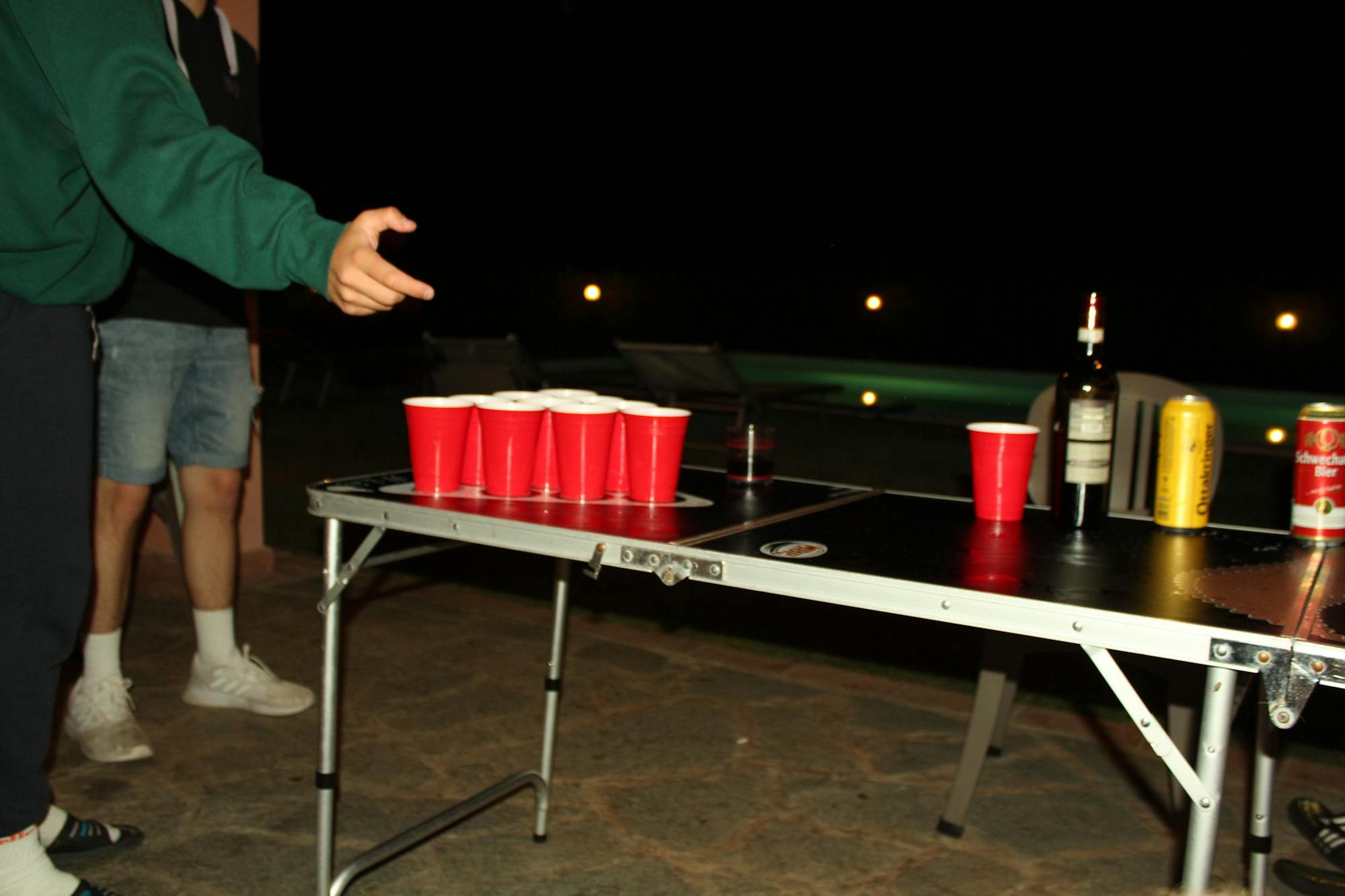 Auf dem Beerpongtisch befinden sich Beerpongbecher sowie Bierdosen und eine Weinflasche. Ein Spieler wirft gerade den Tischtennisball auf die Becher der gegnerischen Mannschaft. 