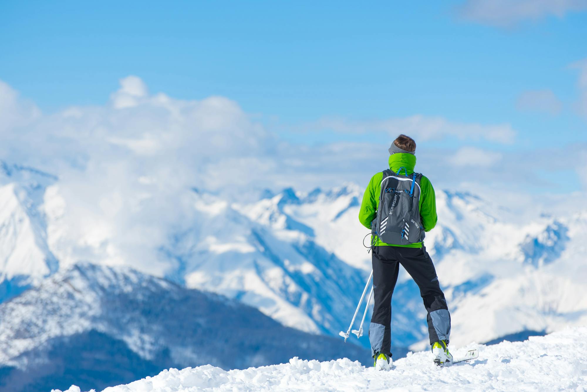 Es ist Winter. Ein Skifahrer befindet sich am Gipfel des Berges und blickt in das schneebedeckte Gipfelpanorama. Die Wolken befinden sich auf gleicher Höhe wie er. 