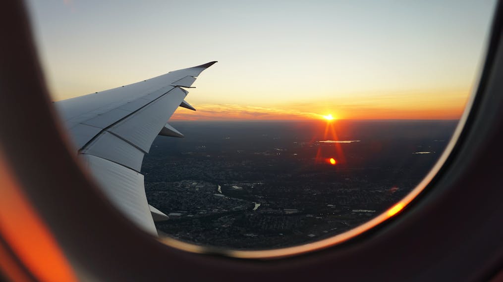 Blick aus dem Fenster eines fliegenden Flugzeugs mit der Aussicht auf eine Großstadt und der untergehenden Sonne. 