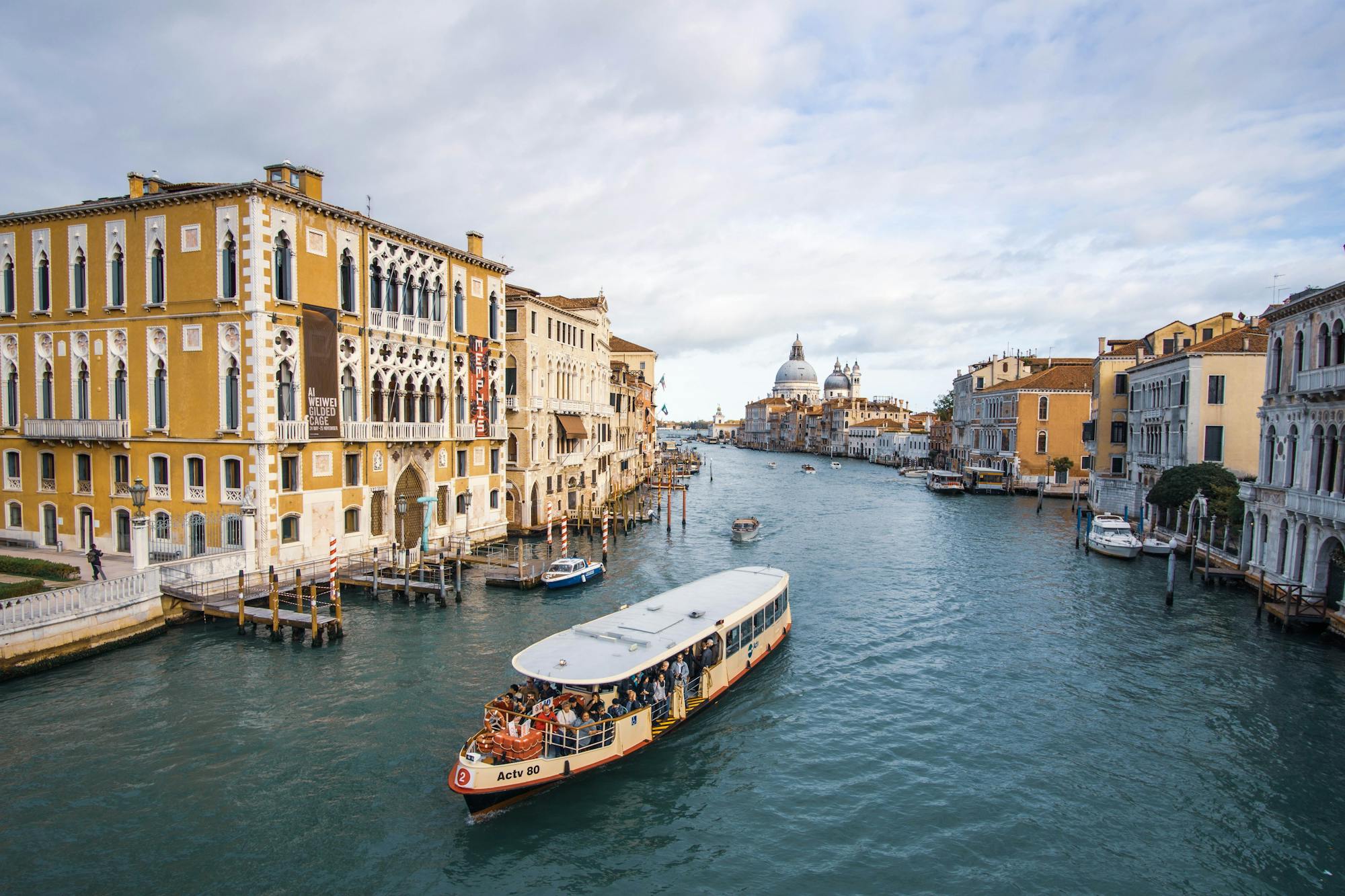 Der Canal Grande, kleine Anlegestellen und schöne italienische Häuser rund herum. Eine Gondelfahrt mit zahlreichen Passagieren ist im Vordergrund des Bildes zu erkennen. 