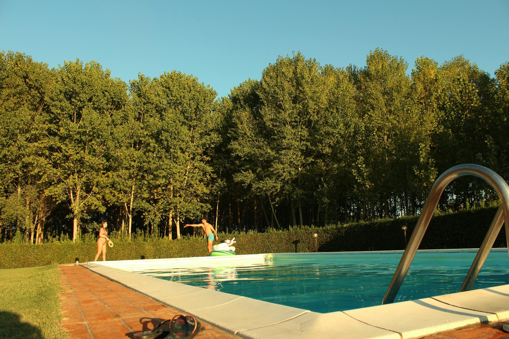 Im Vordergrund befindet sich der Pool, umgeben von grünem Wald. Im Hintergrund spielen zwei Freunde Beach Tennis. 