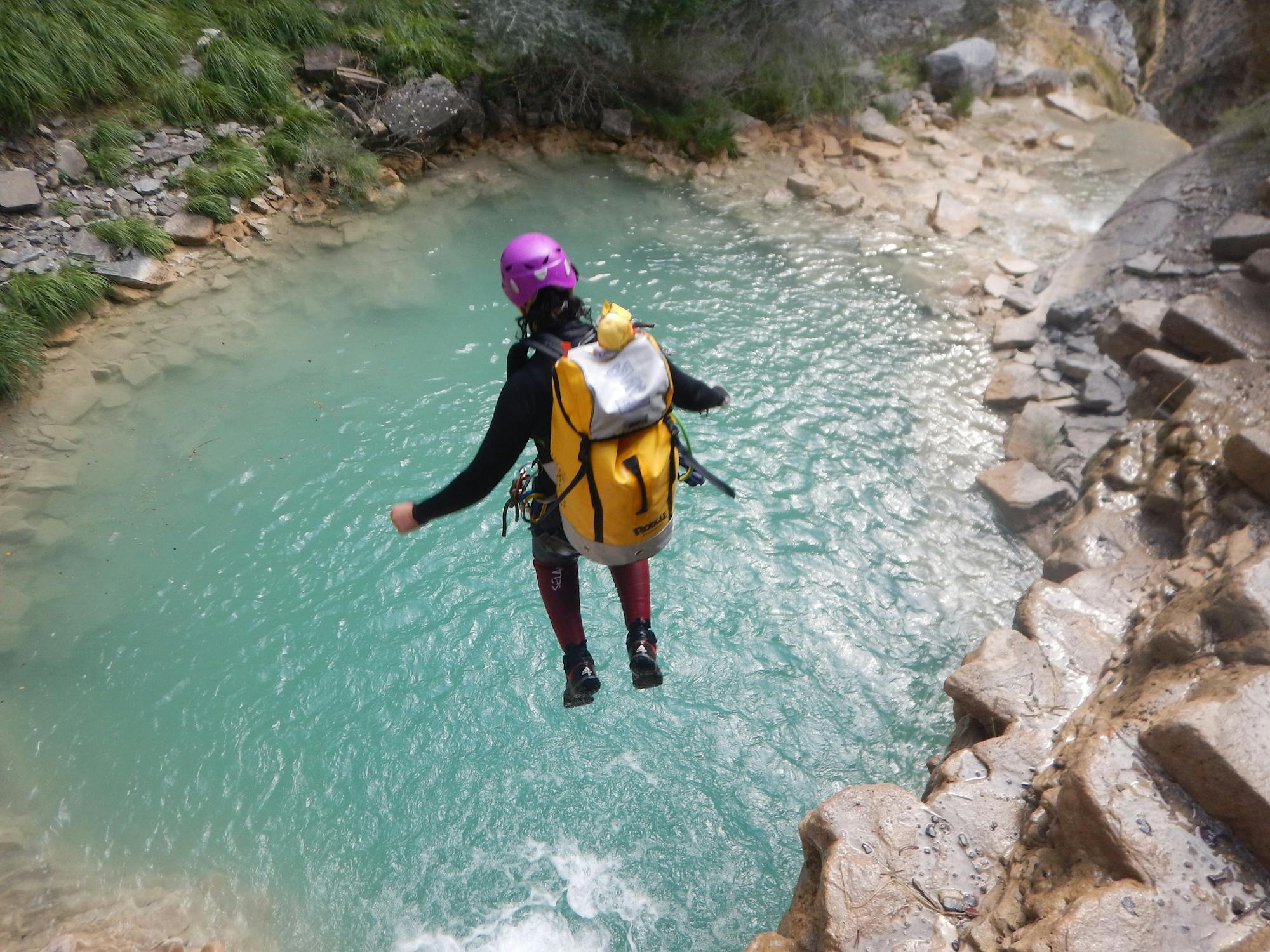 Kobieta w pełnym stroju (plecak, kask, spodnie, kurtka) skacze do błękitnych wód wąwozu. Jest na wycieczce canyoningowej.