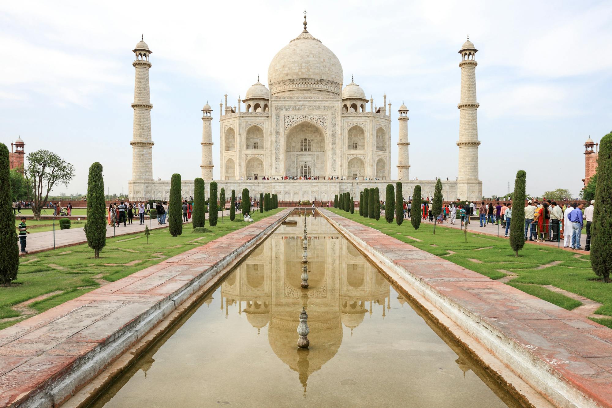 Blick auf den Taj Mahal. Mittig läuft befindet sich ein langgezogener Brunnen, der auf das Mausoleum zuläuft. Drum herum tummeln sich zahlreiche Touristen.