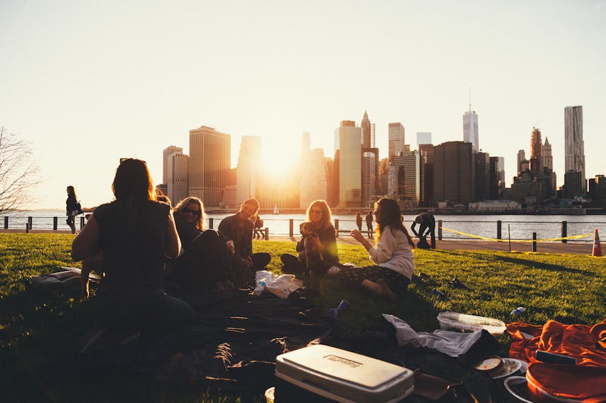Eine Gruppe von Freunden sitzt im Brooklyn Bridge Park und macht ein Picknick. Sie lachen und unterhalten sich. Im Hintergrund sind die hohen Wolkenkratzer und die aufgehende Sonne zu sehen. 