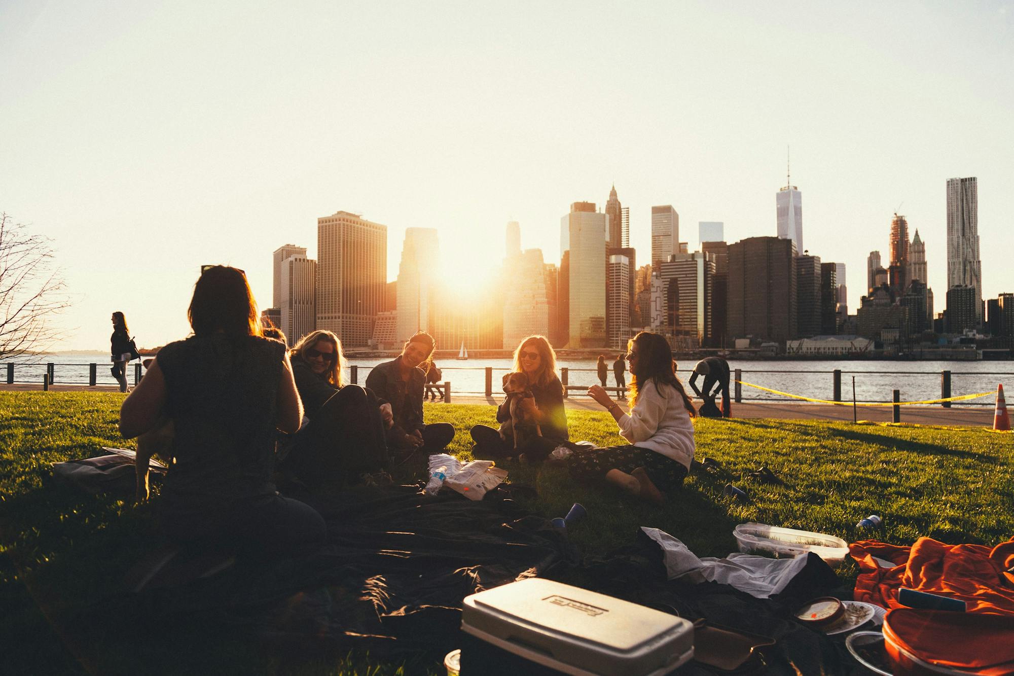 Eine Gruppe von Freunden sitzt im Brooklyn Bridge Park und macht ein Picknick. Sie lachen und unterhalten sich. Im Hintergrund sind die hohen Wolkenkratzer und die aufgehende Sonne zu sehen. 