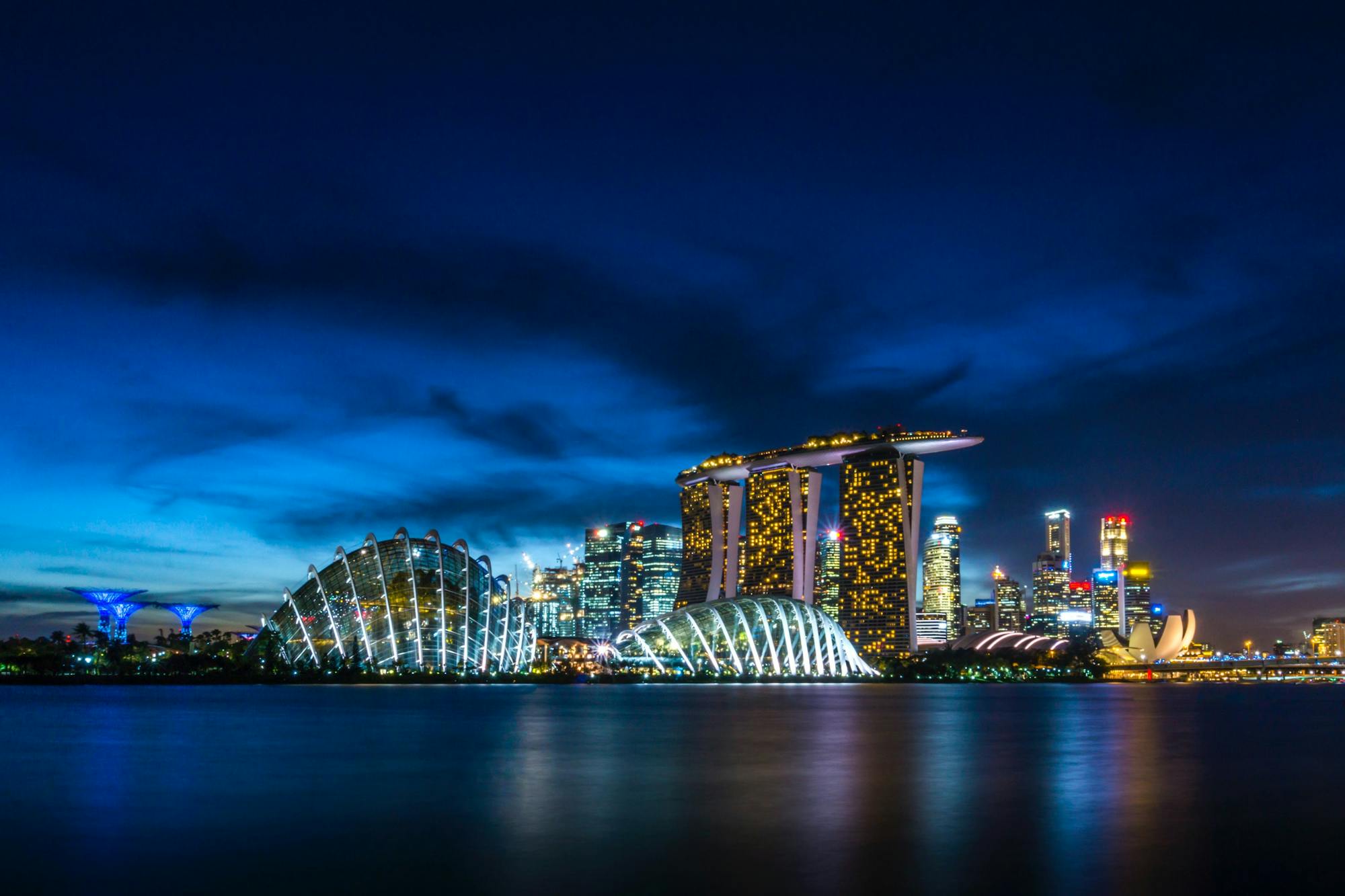 Singapur bei Nacht: beleuchtete Wolkenkratzer und sich im Wasser spiegelnde Lichter. 