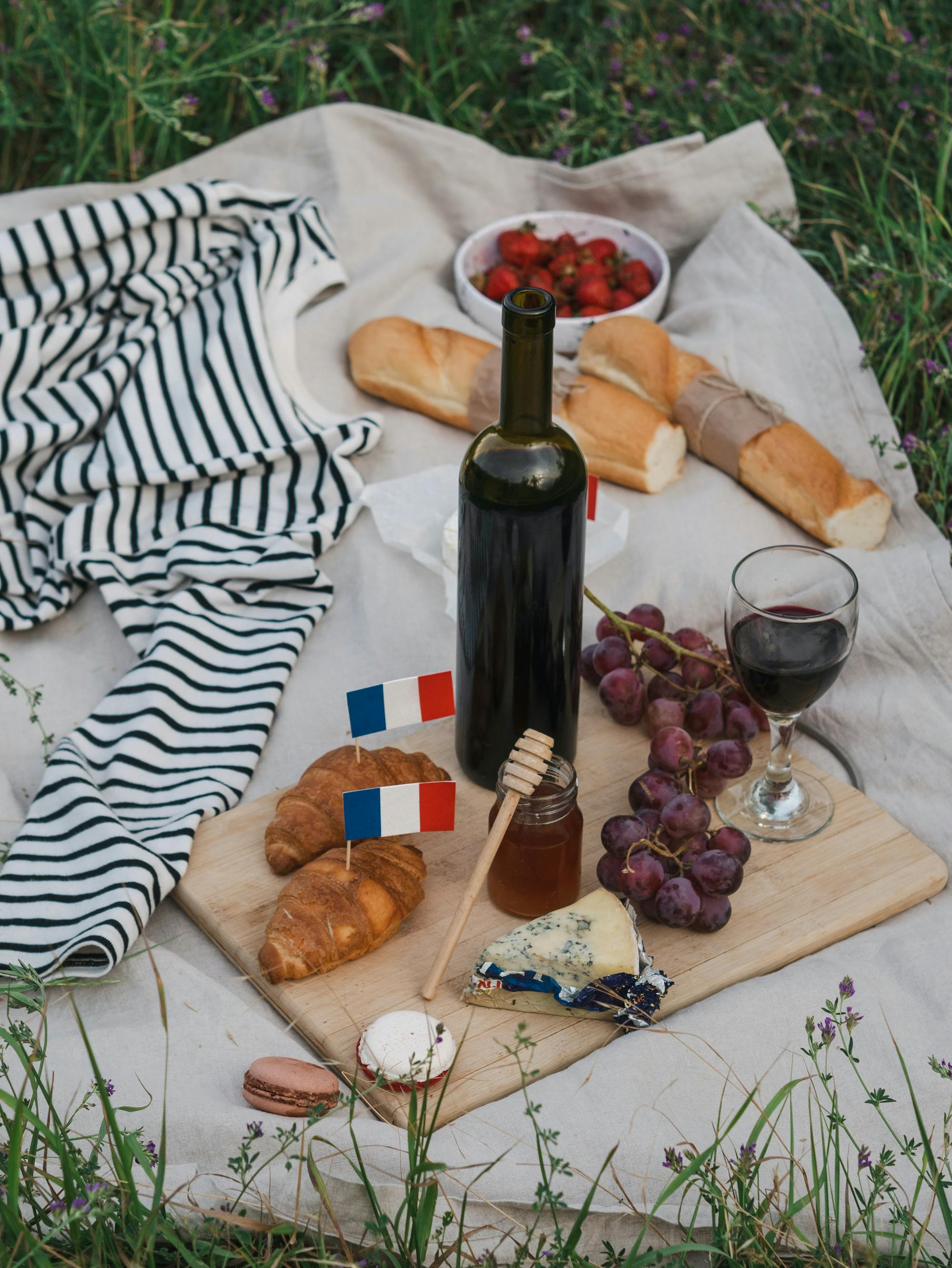 Französisches Picknick auf der Wiese: Auf der Decke liegen schön angerichtet Baguette, Croissant, Wein, Käse und Trauben.