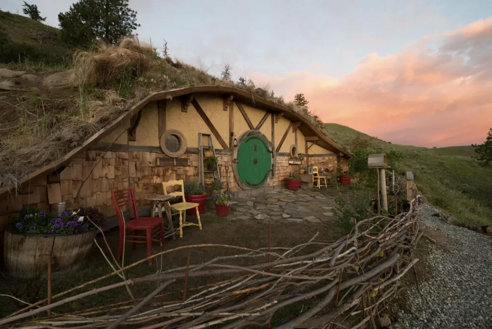 Mały domek hobbita z typowymi okrągłymi drzwiami i uroczymi krzesłami ogrodowymi na przednim podwórku. Domek został wbudowany w wzgórze i otoczony jest cudownym krajobrazem.