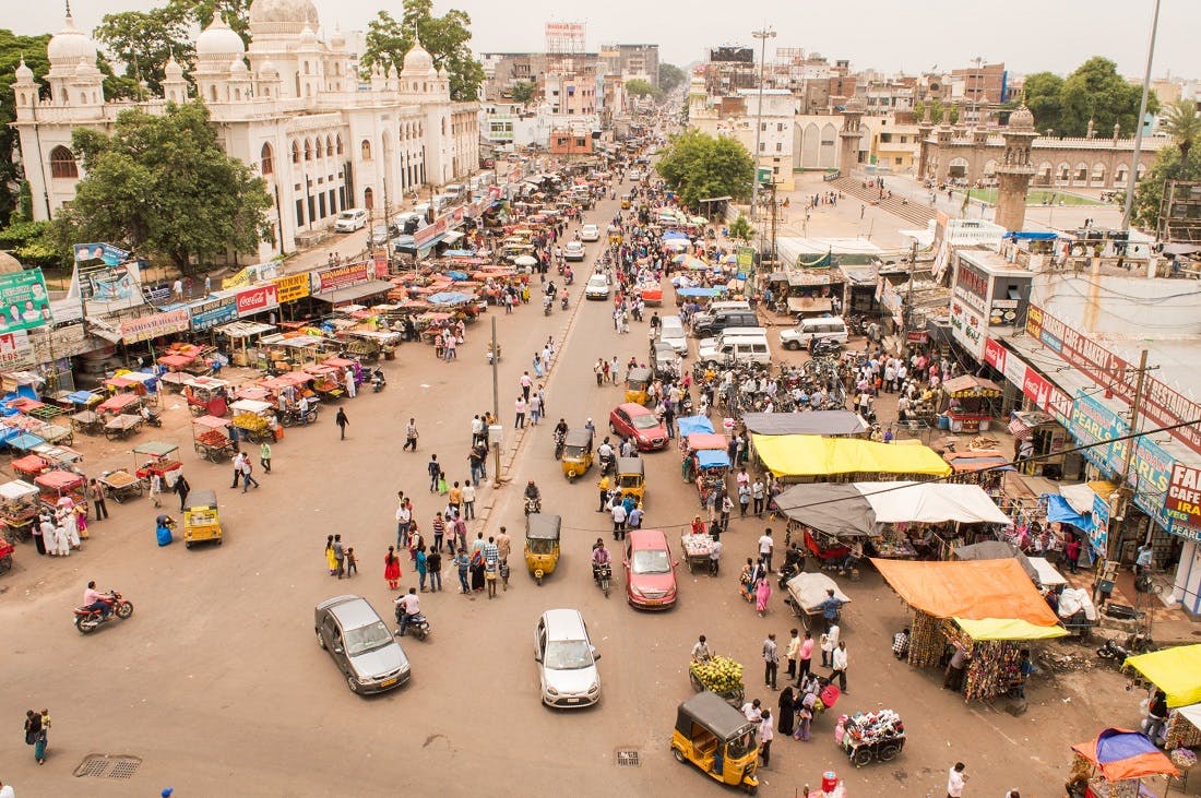 Aus der Vogelperspektive sieht man eine belebte Straße in Indien. Überall sind kleine Stände aufgebaut. Bewohner, Autorikschas, Autos und Motorräder tummeln sich in den Straßen herum. 