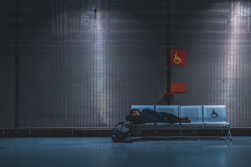 Osoba śpi na ławce przy bramce lotniczej. Jest wieczór, a światła bramki zastępują światło dzienne.