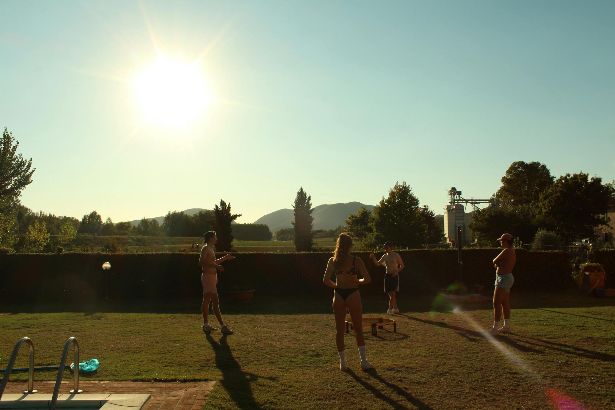 Vier junge Leute spielen gemeinsam Spike Ball (ein Ballspiel). Im Hintergrund ist die strahlende Sonne sowie toskanische Landschaft zu sehen. 