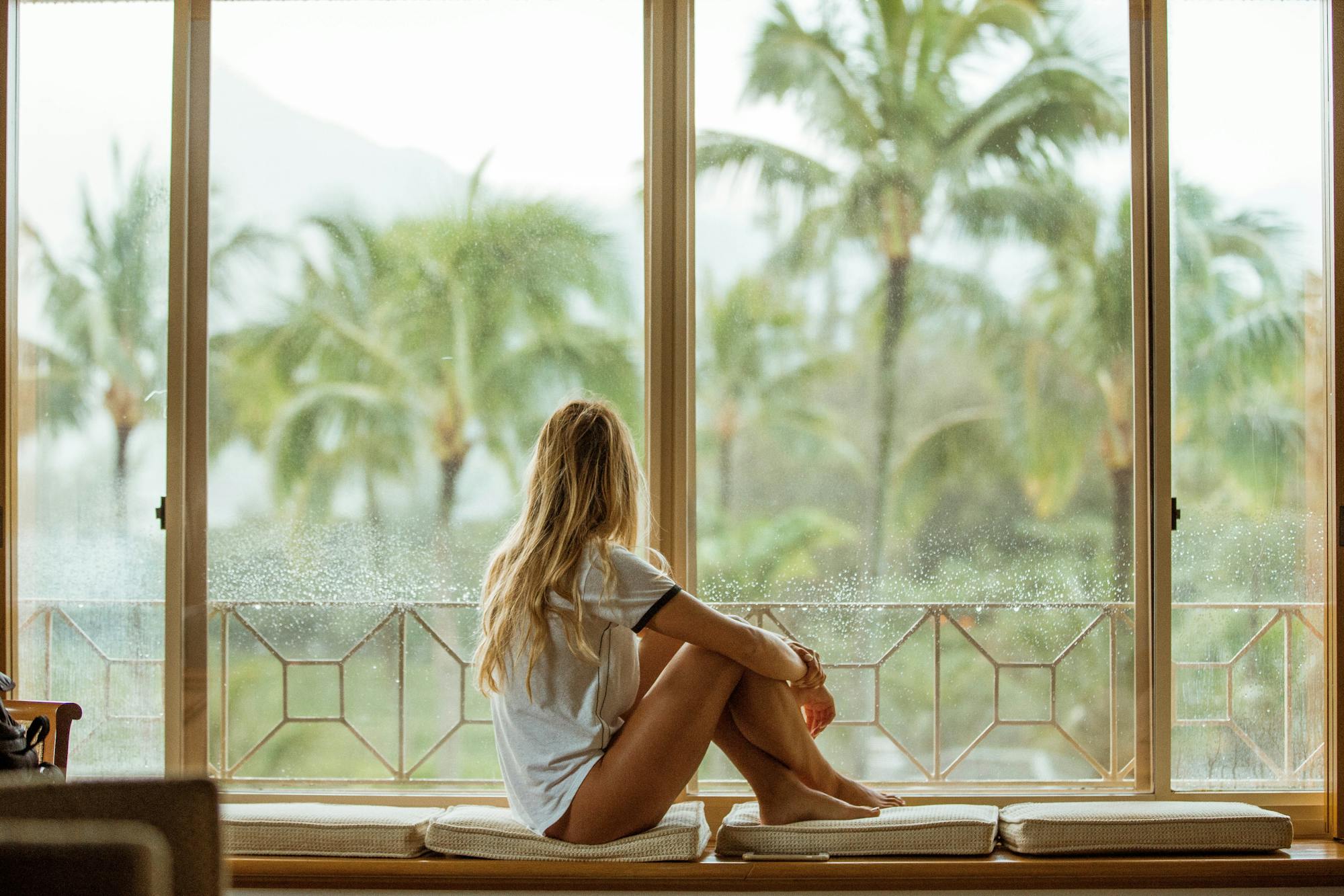 Eine junge Frau sitzt auf der Fensterbank und blickt nach draußen in die Palmen-Landschaft. Das Bild strahlt sommerliche, tropische Gefühle aus.