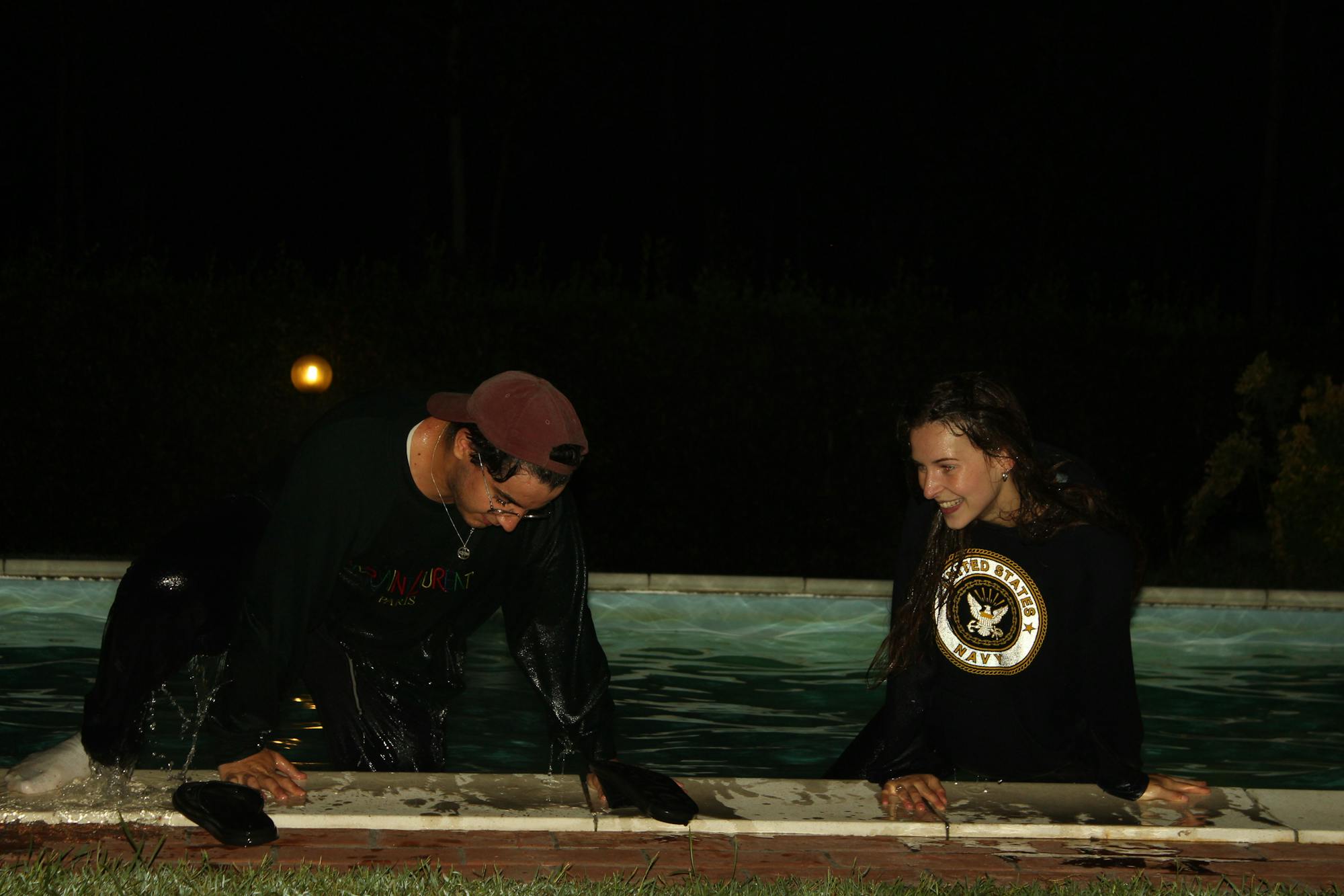 Zwei Freunde stützen sich auf den Poolrand und lachen sich an. Sie sind mit Klamotten nachts in den Pool gesprungen. 