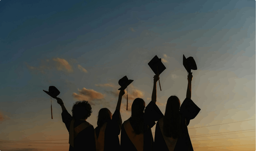 Photograph of girls at sunset raising their graduation caps.