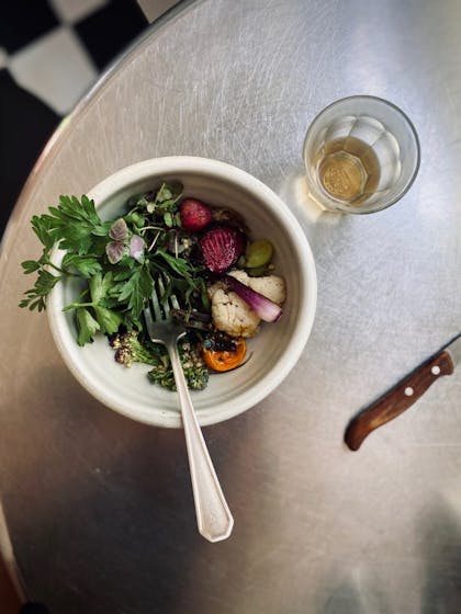 Photo of a bowl of grilled vegetables, a fork, drink, and knife with a wooden blade on a silver table