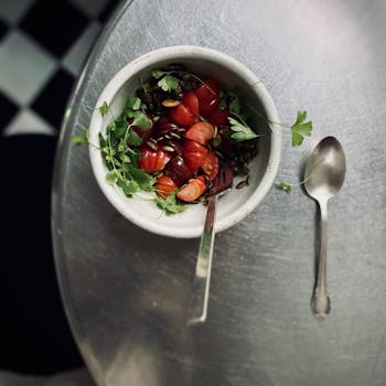 Photo of a bowl of salad wit bright red fruit and silverware on a silver table.