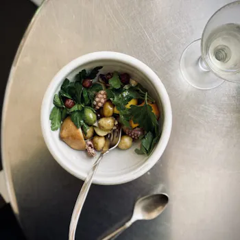 Photo of a bowl of salad, grilled vegetables, and octopus/squid with silverware and a wine glass on a silver table.