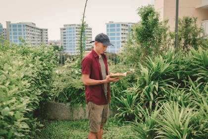 Chris is posing, reading a book amidst pandan plants and others in the rooftop garden