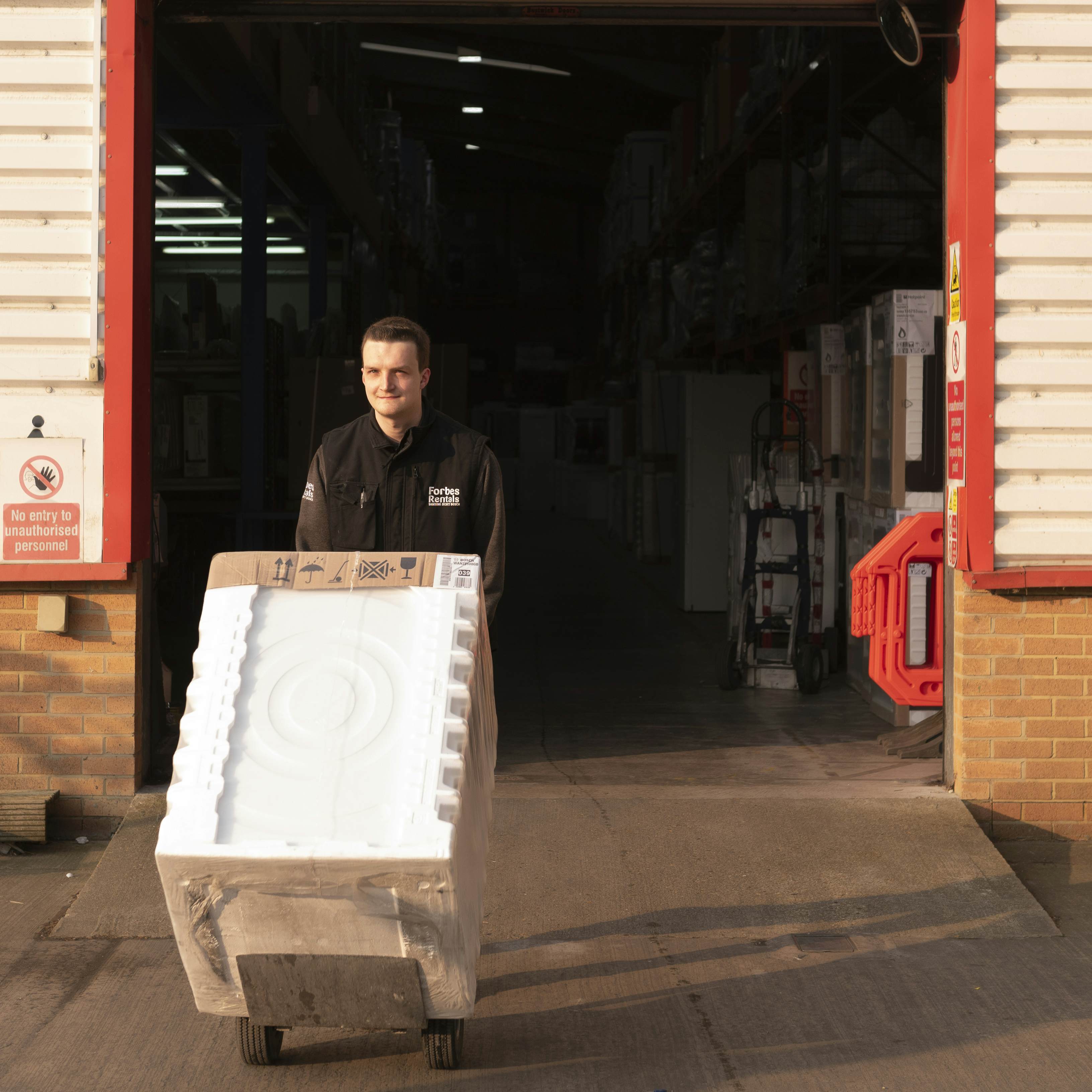 Delivery worker moving a large boxed appliance on a dolly out of a warehouse loading bay.