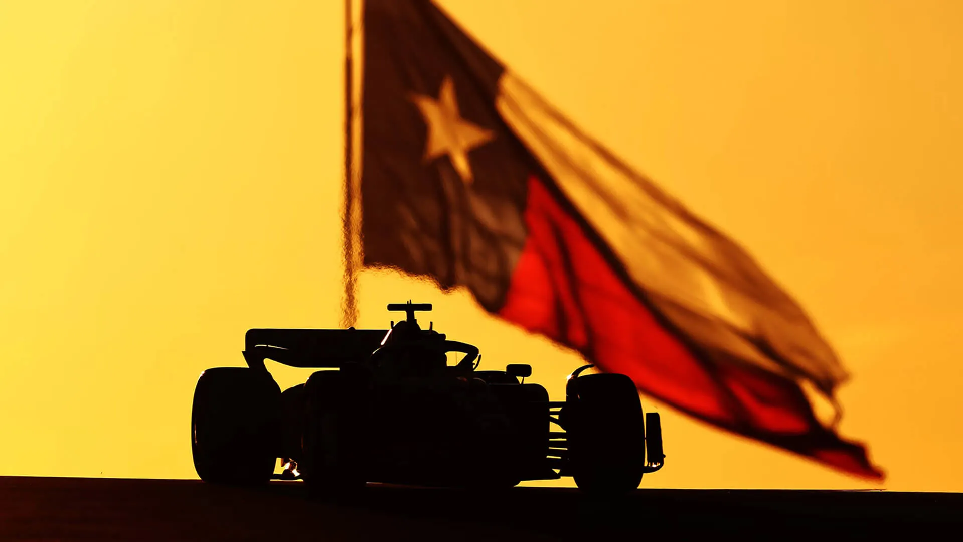 Silhouette of a Formula 1 car against a golden sky, with a Texas flag flying in the background.