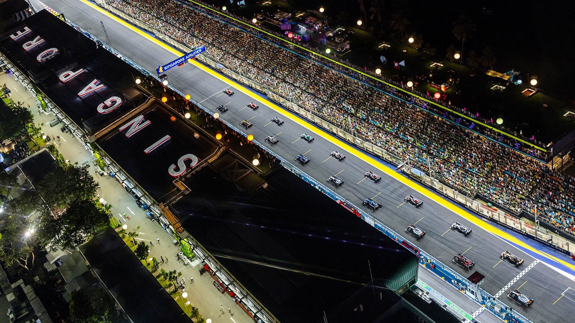 Aerial view of F1 cars on Marina Bay’s floodlit starting grid beside packed grandstands at the Singapore Grand Prix.