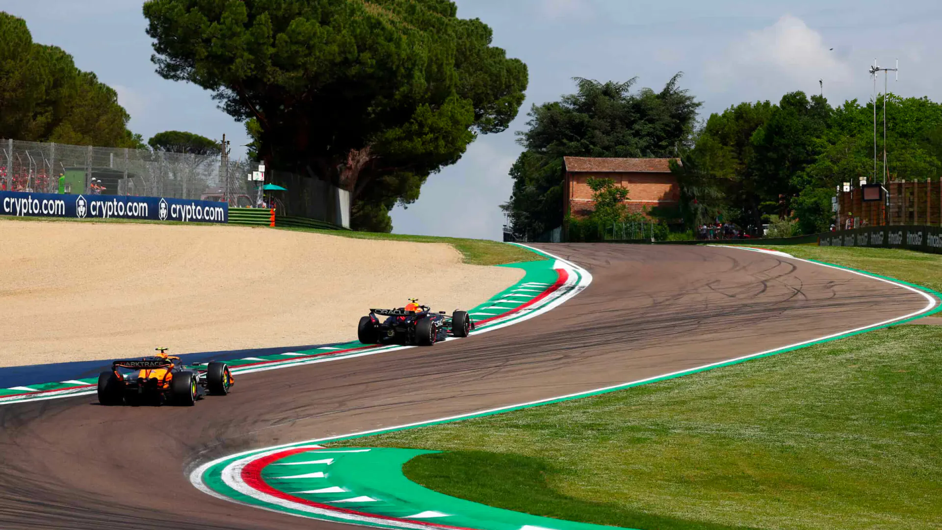 Verstappen and Norris climb the curving section of the Imola track during the 2024 Emilia Romagna GP, with trees and curbs lining the circuit.