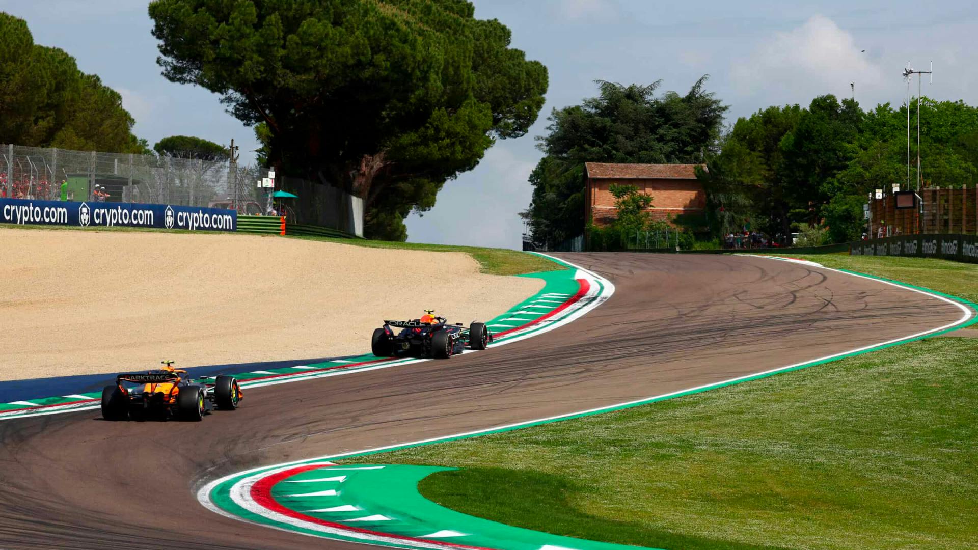 Verstappen and Norris climb the curving section of the Imola track during the 2024 Emilia Romagna GP, with trees and curbs lining the circuit.