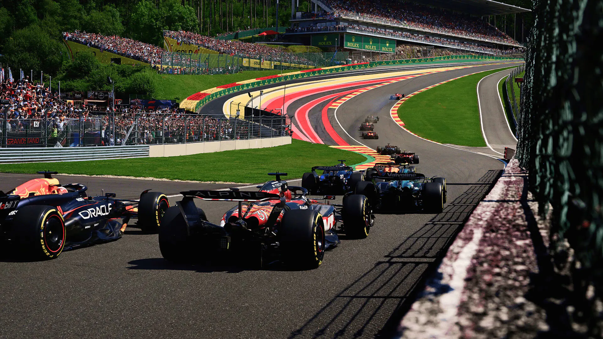 F1 cars race uphill at Spa with colorful run-offs and packed grandstands in view.
