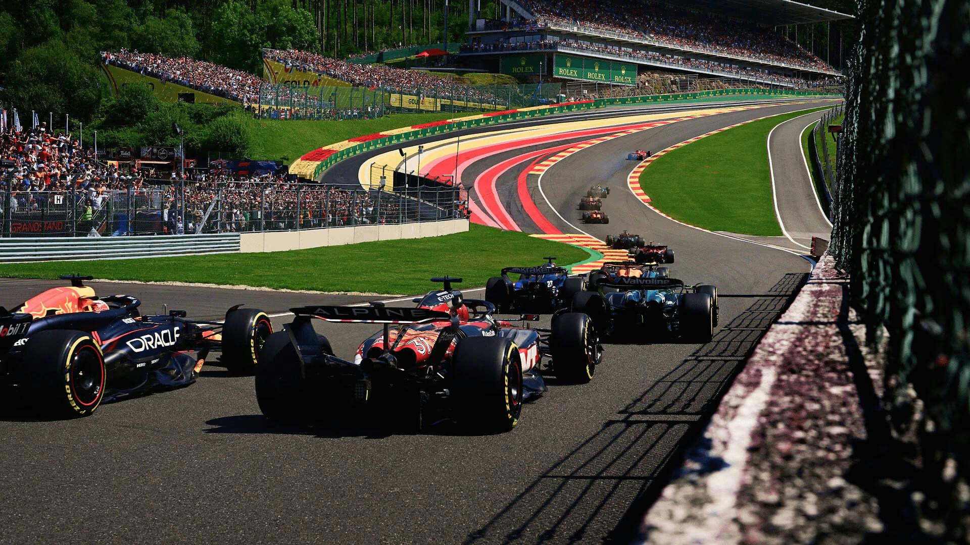 F1 cars race uphill at Spa with colorful run-offs and packed grandstands in view.