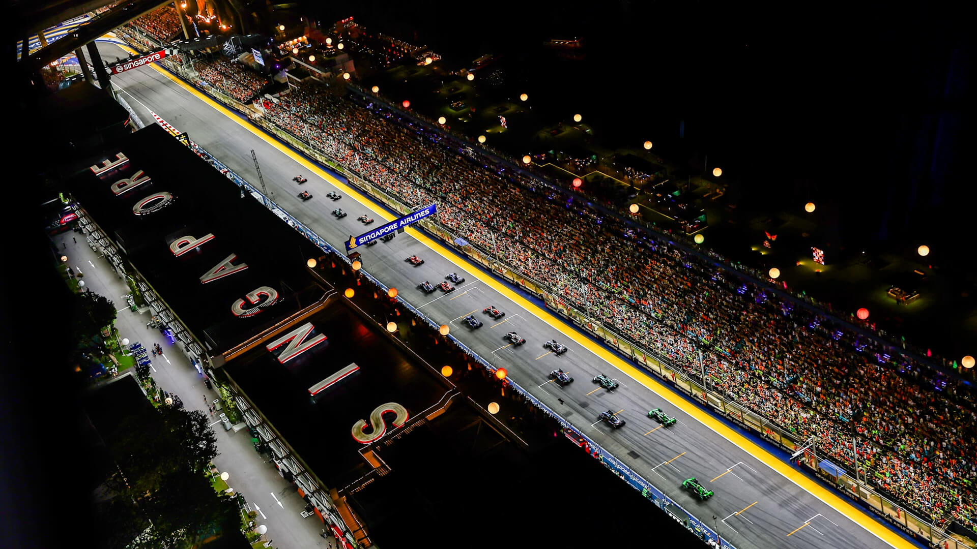 Night-time aerial view of the Singapore Grand Prix track, showing F1 cars driving past grandstands filled with spectators under bright lights.