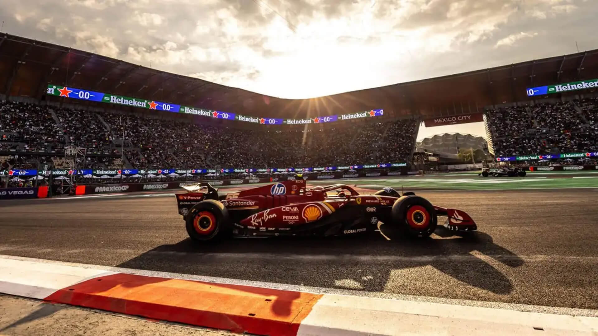 Wide shot of a F1 car driving through the stadium section at the Autódromo Hermanos Rodríguez with packed grandstands at sunset.