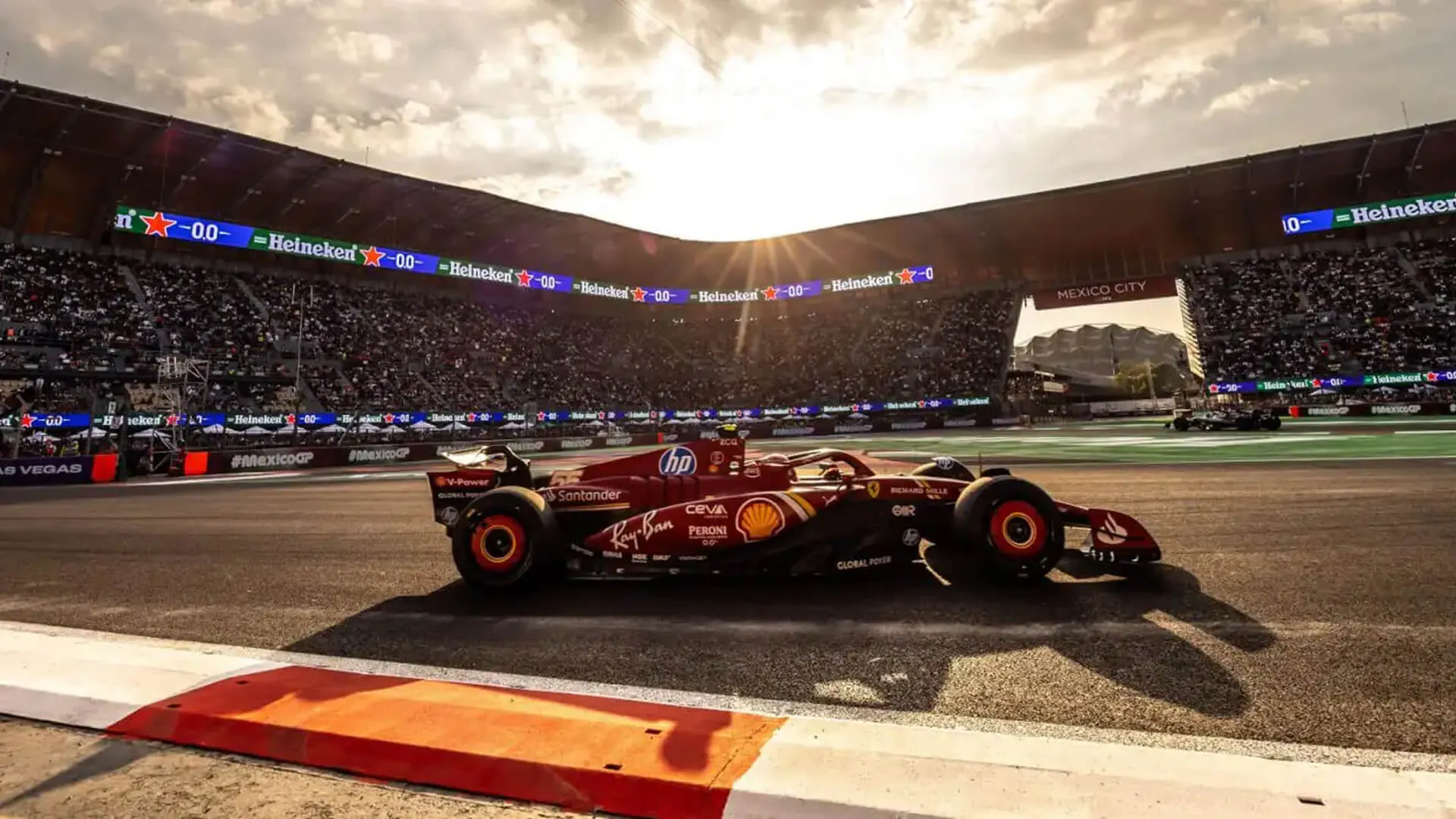 Wide shot of a F1 car driving through the stadium section at the Autódromo Hermanos Rodríguez with packed grandstands at sunset.