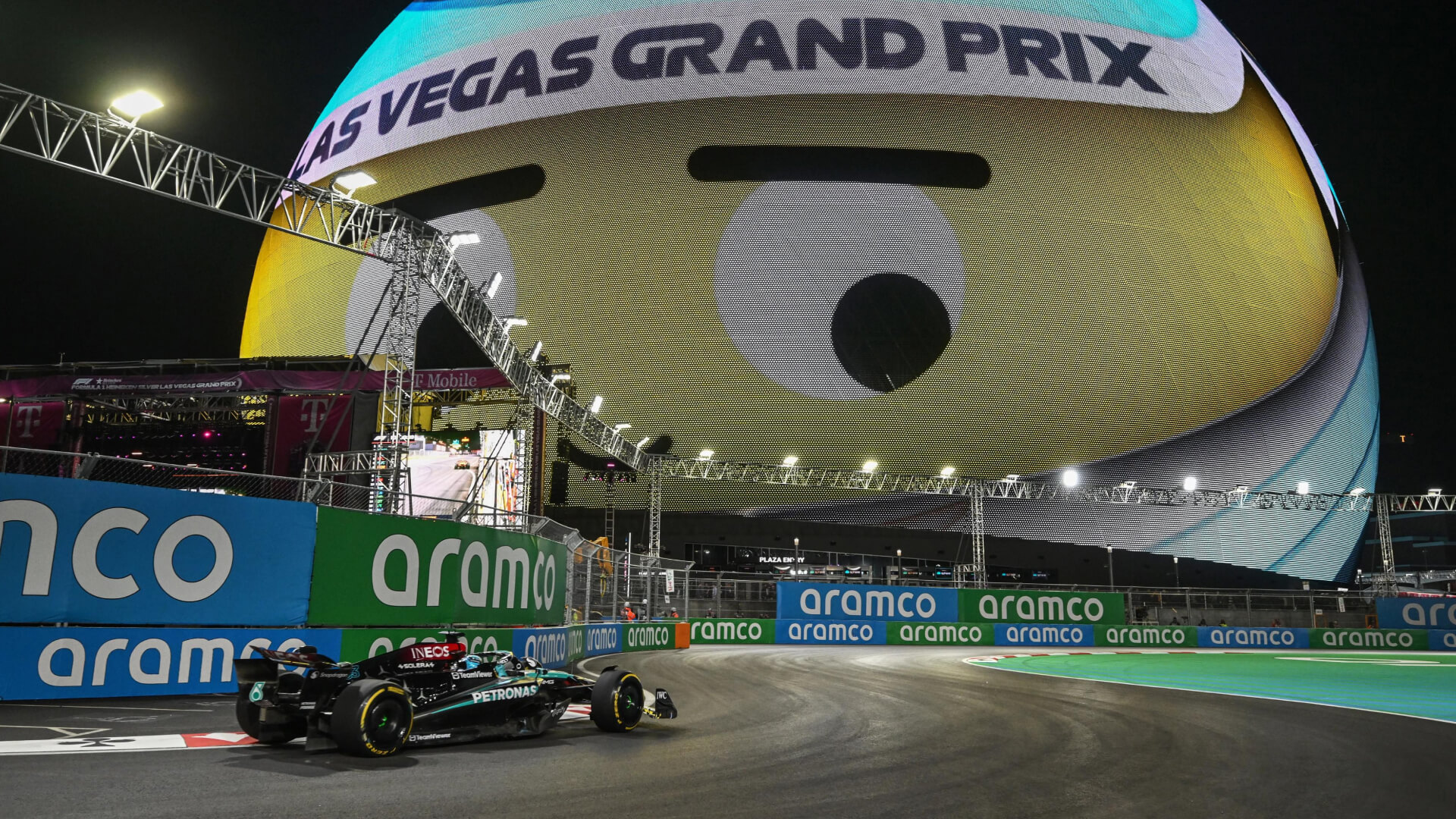 Mercedes F1 car passes under the Las Vegas Sphere displaying a giant winking emoji at night.
