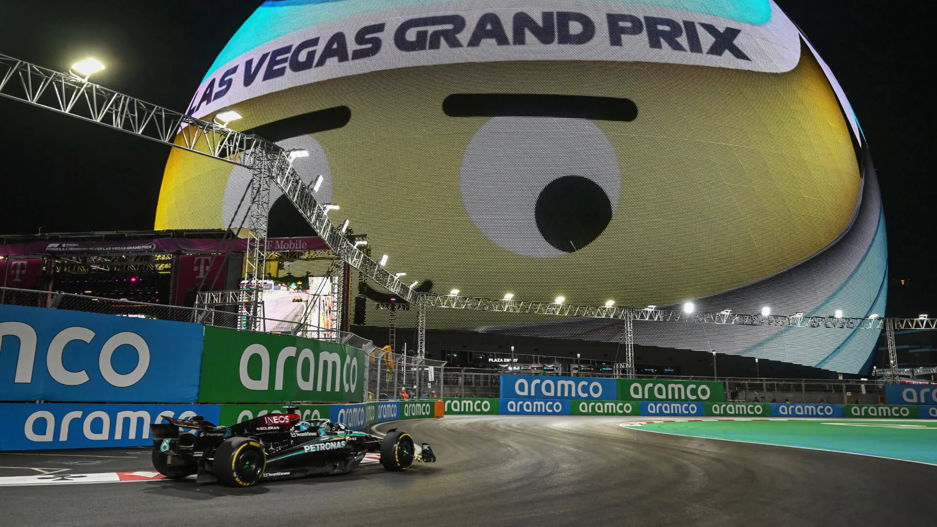 Mercedes F1 car passes under the Las Vegas Sphere displaying a giant winking emoji at night.