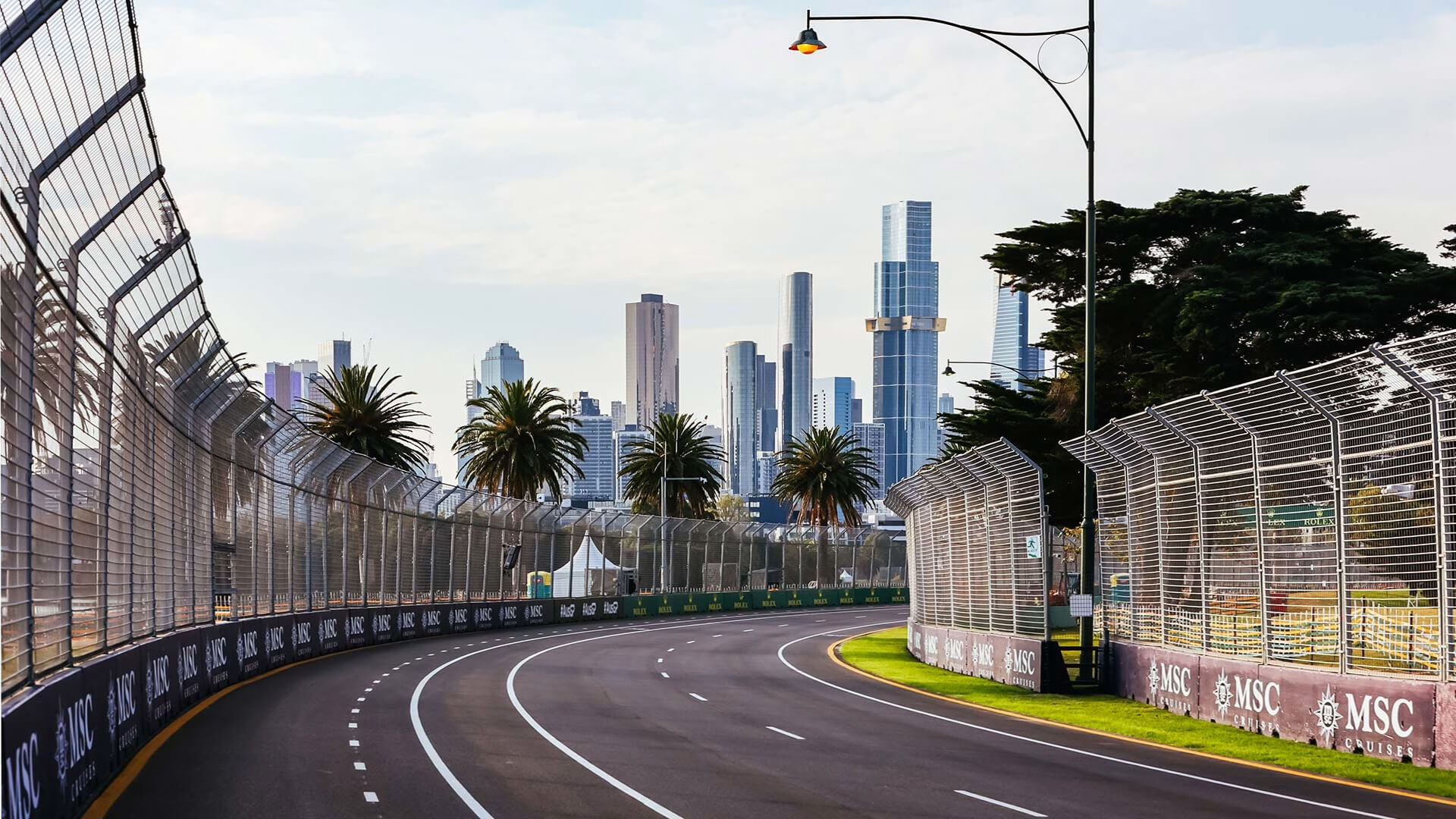 A wide view of the Albert Park Circuit in Melbourne, showing a curved section of track lined. The city skyline and palm trees are visible in the background.