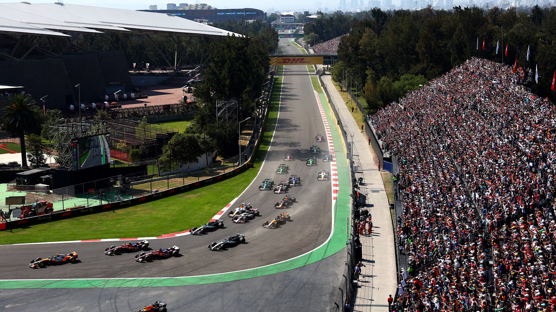 Formula 1 cars approach Turn 1 at the Autódromo Hermanos Rodríguez beside packed grandstands in Mexico City.